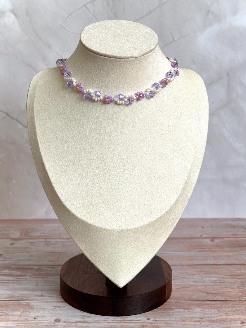 May include: A delicate necklace with a floral design, featuring lavender and white beads. The necklace is displayed on a cream-colored jewelry bust with a dark wooden base. The beads are arranged in a repeating pattern.