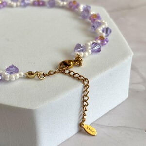 May include: Delicate necklace featuring a strand of lavender and clear crystal beads, interspersed with small white pearls and gold accents. The necklace has a gold-colored chain and clasp, with a small tag that reads "S.Steel".