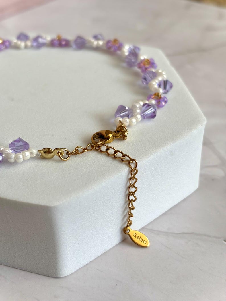 May include: Delicate necklace featuring a strand of lavender and clear crystal beads, interspersed with small white pearls and gold accents. The necklace has a gold-colored chain and clasp, with a small tag that reads "S.Steel".