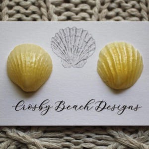 May include: A pair of yellow seashell earrings on a white card with a seashell illustration and the text "Crosby Beach Designs". The earrings are set against a textured, neutral-toned background.