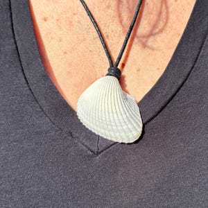 May include: A white seashell pendant hangs from a black cord necklace. The shell is a light, natural colour with a ridged texture. The necklace is worn against a dark grey v-neck shirt.