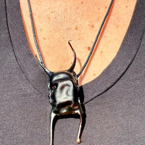 May include: A black pendant necklace with a unique, abstract design. The pendant has a textured, irregular shape with two upward-curving elements and two downward-hanging legs with gold-colored tips. The necklace cord is black.