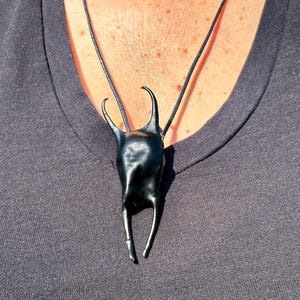 Puede incluir: Un collar con colgante negro de forma estilizada, posiblemente inspirado en la vida marina. El colgante cuelga de un fino cordón negro, sobre una camiseta gris oscuro. El colgante tiene dos cuernos curvos y dos patas rectas.