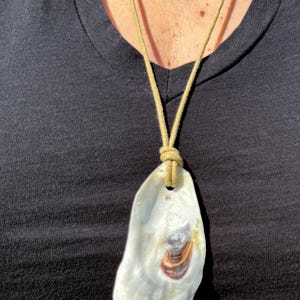 May include: A handmade necklace featuring a large, pearly white oyster shell pendant with brown and tan markings. The shell is suspended from a beige cord, creating a natural and unique accessory.