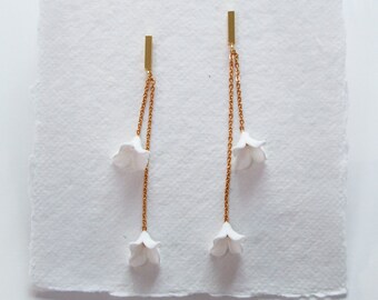 May include: A pair of gold chain earrings with white flower accents. The earrings are displayed on a white card with the word "bespoke" written in black.