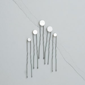 May include: Six silver hair clips with round, white, reflective tops. The clips are arranged in a staggered pattern.