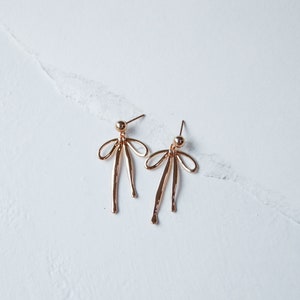 Pode incluir: Um par de brincos dourados com um design de laço. Os brincos são feitos de metal e têm um estilo simples e elegante.