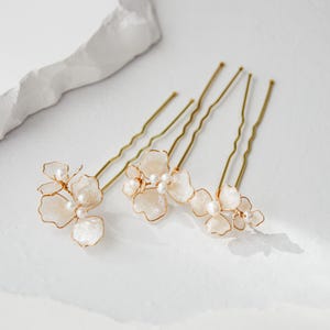 May include: Four gold-toned hair pins with delicate, white flower accents and small pearl embellishments. The floral designs are clustered on the pins, creating a refined and elegant look. Ideal for weddings or special occasions.