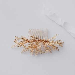 May include: A gold-coloured hair comb with a delicate leaf design. The comb has a series of thin, evenly spaced tines, with the decorative leaves extending outwards. The leaves are detailed and add a touch of elegance to the hair accessory.