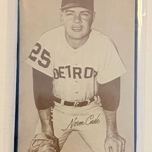 May include: Sepia-toned baseball card featuring a player in a Detroit Tigers uniform. The player wears a cap with a 'B' logo and the word 'DETROIT' on the jersey. The number '25' is visible on the sleeve, and the name 'Norm Cash' is printed on the pants.