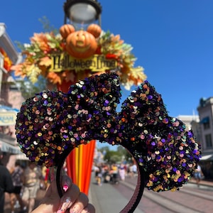 May include: Black and iridescent sequined Minnie Mouse ears headband with a pink velvet headband. The headband is in front of a sign that says "Halloween Time".