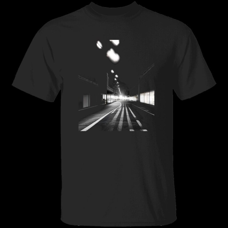 Pode incluir: Uma t-shirt preta de manga curta com uma impress&atilde;o gr&aacute;fica quadrada a preto e branco centrada, representando uma rua da cidade deserta &agrave; noite. A imagem mostra uma estrada com marca&ccedil;&otilde;es de faixa, montras iluminadas e candeeiros.
