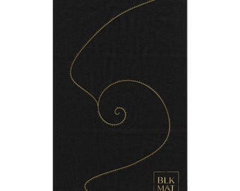 Op de afbeelding: Een rechthoekige zwarte handdoek met een goudkleurige spiraalafbeelding bestaande uit kleine streepjes. In de rechterbenedenhoek staat een goudkleurig vierkant kader met de tekst BLK MAT in een strak, schreefloos lettertype.