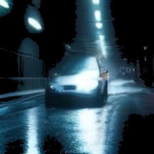 May include: A silver car driving through a dark, wet tunnel illuminated by overhead lights. The vehicle's headlights are bright, casting a glow on the reflective road surface, while the surrounding tunnel walls remain in deep shadow.