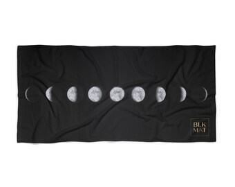 May include: A rectangular black towel featuring a horizontal sequence of moon phases in shades of grey and white. The bottom right corner displays a small square logo with the gold text BLK MAT. The fabric has a soft, folded texture throughout.