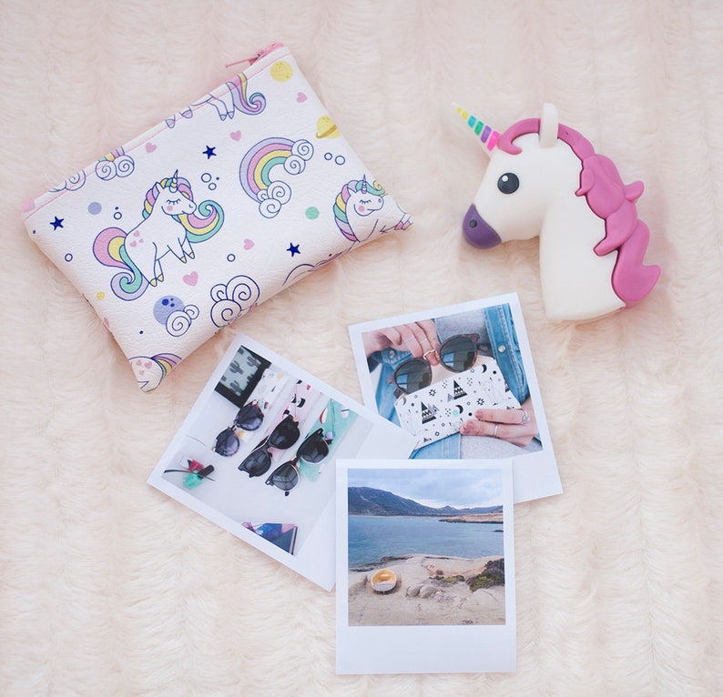 K&ouml;nnte beinhalten: Eine wei&szlig;e Einhorn-Geldb&ouml;rse mit Rei&szlig;verschluss, mit Einh&ouml;rnern, Regenbogen und Sternen. Ein wei&szlig;es Einhorn-Nachtlicht mit rosa M&auml;hne und Horn. Polaroids mit Sonnenbrillen und einer malerischen Aussicht.