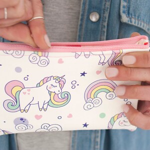 K&ouml;nnte beinhalten: Eine wei&szlig;e Rei&szlig;verschlusstasche mit rosa Rei&szlig;verschluss und Besatz. Die Tasche ist mit einem sich wiederholenden Muster aus Einh&ouml;rnern, Regenbogen, Sternen und Herzen in Pastellfarben verziert. Die Tasche wird ge&ouml;ffnet gehalten.