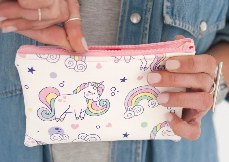 K&ouml;nnte beinhalten: Eine wei&szlig;e Rei&szlig;verschlusstasche mit rosa Rei&szlig;verschluss und Besatz. Die Tasche ist mit einem sich wiederholenden Muster aus Einh&ouml;rnern, Regenbogen, Sternen und Herzen in Pastellfarben verziert. Die Tasche wird ge&ouml;ffnet gehalten.