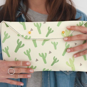 May include: Cream-colored clutch with a repeating pattern of green cacti and small red dots. The clutch has a flap closure secured by two gold-colored studs. The edges are stitched with black thread.