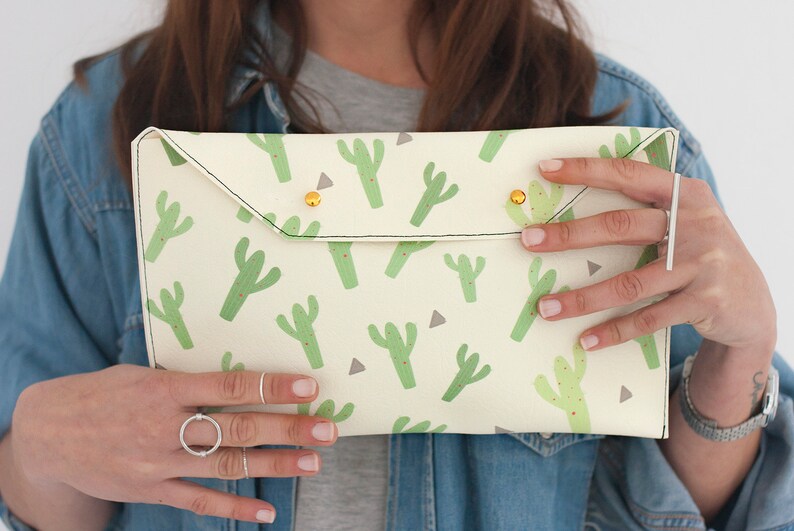 May include: Cream-colored clutch with a repeating pattern of green cacti and small red dots. The clutch has a flap closure secured by two gold-colored studs. The edges are stitched with black thread.