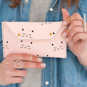 May include: A light pink clutch with a cat face pattern. The clutch features a flap closure with two gold-colored rivets. The cat faces are black and white, with closed eyes and whiskers. The clutch is held by a person wearing a denim shirt.