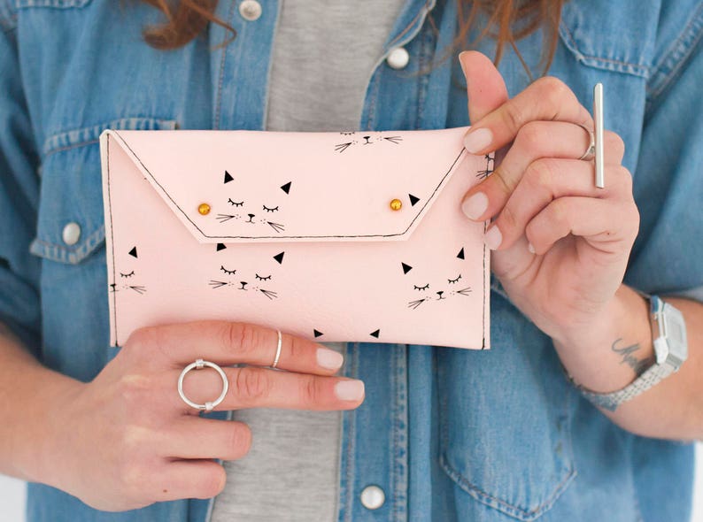 May include: A light pink clutch with a cat face pattern. The clutch features a flap closure with two gold-colored rivets. The cat faces are black and white, with closed eyes and whiskers. The clutch is held by a person wearing a denim shirt.