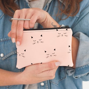 May include: A light pink zippered pouch featuring a repeating pattern of black cat faces with closed eyes and whiskers. The pouch has a black zipper and is held in front of a denim jacket.