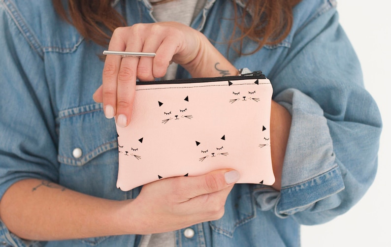 May include: A light pink zippered pouch featuring a repeating pattern of black cat faces with closed eyes and whiskers. The pouch has a black zipper and is held in front of a denim jacket.