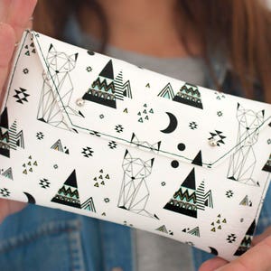 May include: White clutch with a geometric fox and mountain pattern. The design includes black triangles, a crescent moon, and small geometric shapes. The clutch has a flap closure with two silver snaps.