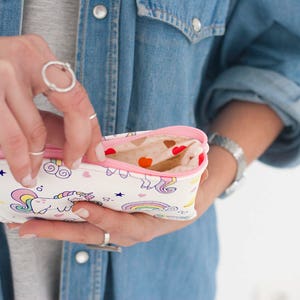 K&ouml;nnte beinhalten: Ein wei&szlig;es Federm&auml;ppchen mit rosa Rei&szlig;verschluss und Futter, verziert mit einem Einhorn, Regenbogen und Sternen. Das M&auml;ppchen ist ge&ouml;ffnet und zeigt ein gemustertes Inneres mit Herzformen.