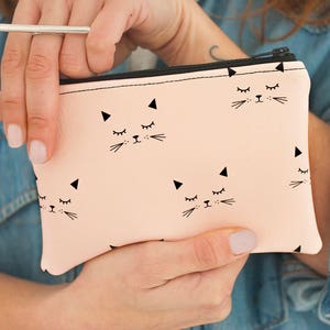 May include: A light pink zippered pouch featuring a repeating pattern of black cat faces with closed eyes and triangular ears. The pouch has a black zipper and is held by a person wearing a denim shirt.