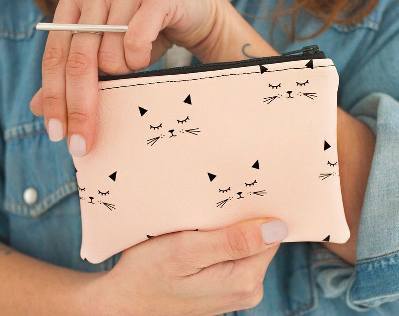 May include: A light pink zippered pouch featuring a repeating pattern of black cat faces with closed eyes and triangular ears. The pouch has a black zipper and is held by a person wearing a denim shirt.