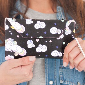 May include: Black clutch with a whimsical unicorn and cloud pattern. The clutch features a flap closure with pink stitching. The design includes white unicorns, clouds, stars, and hearts on a black background.