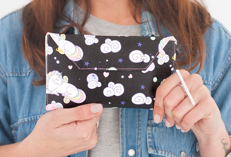 May include: Black clutch with a whimsical unicorn and cloud pattern. The clutch features a flap closure with pink stitching. The design includes white unicorns, clouds, stars, and hearts on a black background.