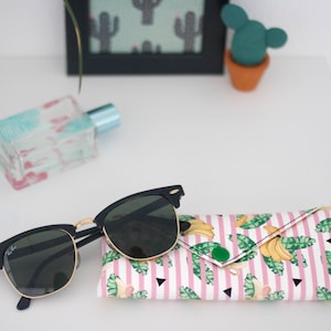 Puede incluir: Gafas de sol negras con detalles dorados sobre un estuche estampado. El estuche presenta un fondo a rayas blancas y rosas con diseños de plátanos y hojas. Un frasco de perfume y decoración de cactus en el fondo.