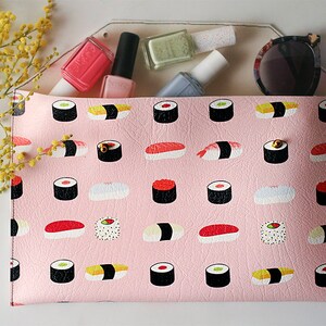 May include: A pink clutch with a sushi pattern, holding nail polish bottles and sunglasses. The clutch is decorated with illustrations of various sushi rolls. A sprig of yellow flowers is in the background.