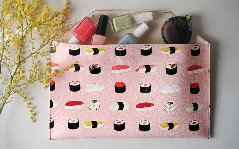 May include: A pink clutch with a sushi pattern, holding nail polish bottles and sunglasses. The clutch is decorated with illustrations of various sushi rolls. A sprig of yellow flowers is in the background.