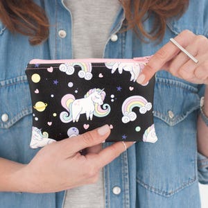 K&ouml;nnte beinhalten: Schwarze Rei&szlig;verschlusstasche mit einem Einhorn- und Regenbogenmuster. Das Design umfasst wei&szlig;e Einh&ouml;rner, bunte Regenbogen, Planeten und Sterne auf schwarzem Hintergrund. Die Tasche hat einen rosa Rei&szlig;verschluss und wird von einer Person gehalten.