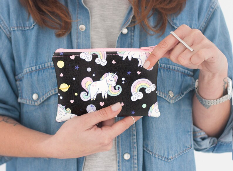 K&ouml;nnte beinhalten: Schwarze Rei&szlig;verschlusstasche mit einem Einhorn- und Regenbogenmuster. Das Design umfasst wei&szlig;e Einh&ouml;rner, bunte Regenbogen, Planeten und Sterne auf schwarzem Hintergrund. Die Tasche hat einen rosa Rei&szlig;verschluss und wird von einer Person gehalten.