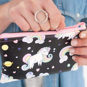 K&ouml;nnte beinhalten: Schwarze Rei&szlig;verschlusstasche mit rosa Rei&szlig;verschluss und Futter. Die Tasche hat ein skurriles Design mit Einh&ouml;rnern, Regenbogen, Planeten und Sternen. Die Tasche wird ge&ouml;ffnet gehalten und gibt den Blick ins Innere frei.