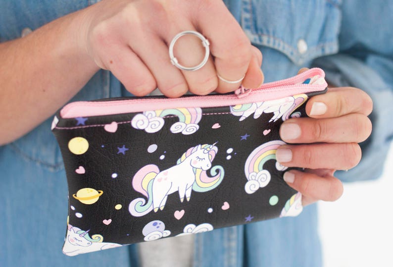 K&ouml;nnte beinhalten: Schwarze Rei&szlig;verschlusstasche mit rosa Rei&szlig;verschluss und Futter. Die Tasche hat ein skurriles Design mit Einh&ouml;rnern, Regenbogen, Planeten und Sternen. Die Tasche wird ge&ouml;ffnet gehalten und gibt den Blick ins Innere frei.