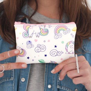 K&ouml;nnte beinhalten: Eine wei&szlig;e Rei&szlig;verschlusstasche mit einem Muster aus Einh&ouml;rnern, Regenbogen und Wolken in Pastellfarben. Die Tasche hat einen rosa Rei&szlig;verschluss und wird vor einer Jeansjacke gehalten. Das Design umfasst Sterne und Planeten.