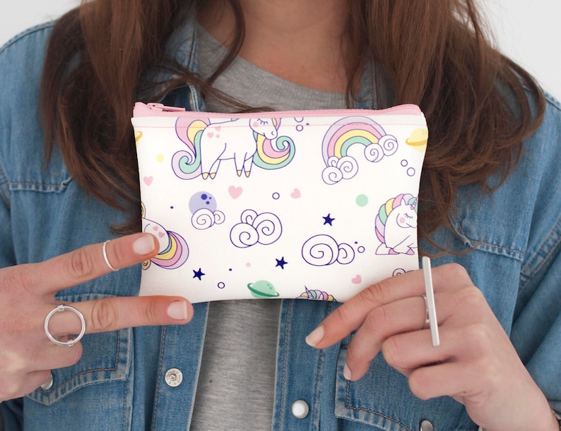 K&ouml;nnte beinhalten: Eine wei&szlig;e Rei&szlig;verschlusstasche mit einem Muster aus Einh&ouml;rnern, Regenbogen und Wolken in Pastellfarben. Die Tasche hat einen rosa Rei&szlig;verschluss und wird vor einer Jeansjacke gehalten. Das Design umfasst Sterne und Planeten.