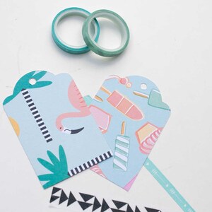 K&ouml;nnte beinhalten: Zwei Rollen t&uuml;rkisfarbenes Washi-Tape und zwei hellblaue Geschenkanh&auml;nger mit Flamingo- und Eis-Design. Ein schwarz-wei&szlig; gemustertes Klebeband und ein t&uuml;rkisfarbenes Pfeilmusterband sind ebenfalls sichtbar. Die Anh&auml;nger haben eine gezackte Oberseite.