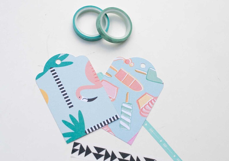 K&ouml;nnte beinhalten: Zwei Rollen t&uuml;rkisfarbenes Washi-Tape und zwei hellblaue Geschenkanh&auml;nger mit Flamingo- und Eis-Design. Ein schwarz-wei&szlig; gemustertes Klebeband und ein t&uuml;rkisfarbenes Pfeilmusterband sind ebenfalls sichtbar. Die Anh&auml;nger haben eine gezackte Oberseite.
