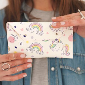 May include: A white clutch with a whimsical design featuring rainbows, unicorns, stars, and pink planets. The clutch has a flap closure with a snap and is held by a person wearing a denim jacket.