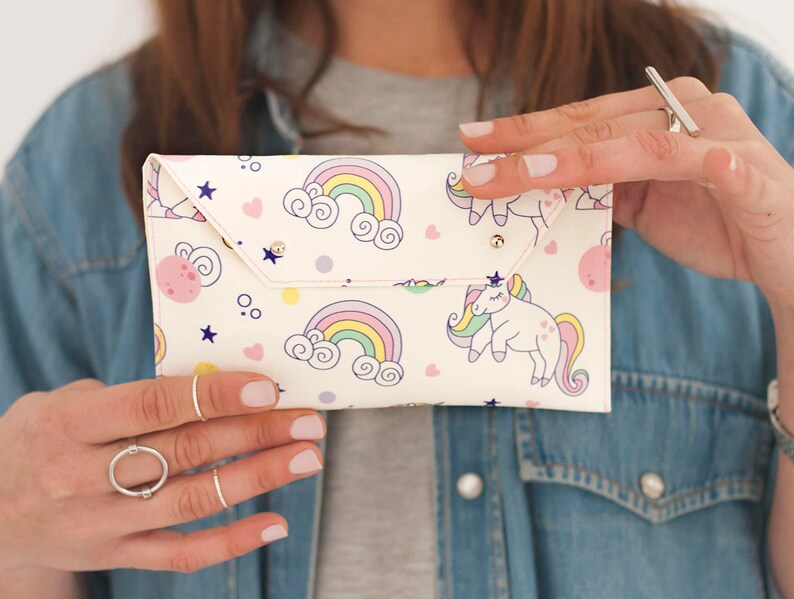 May include: A white clutch with a whimsical design featuring rainbows, unicorns, stars, and pink planets. The clutch has a flap closure with a snap and is held by a person wearing a denim jacket.