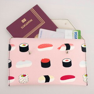 May include: A pink travel wallet with a sushi pattern, holding a passport and credit cards. The wallet features a repeating design of various sushi types. The passport is burgundy with the word "PASAPORTE" printed on it.
