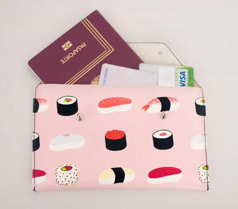 May include: A pink travel wallet with a sushi pattern, holding a passport and credit cards. The wallet features a repeating design of various sushi types. The passport is burgundy with the word "PASAPORTE" printed on it.