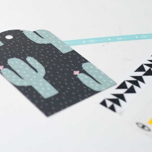 K&ouml;nnte beinhalten: Ein schwarzes Geschenk-Etikett mit einem Kaktus-Design in Hellgr&uuml;n und Rosa. Dekoratives Washi-Tape mit geometrischen Mustern in Schwarz und Wei&szlig; sowie Gelb und Wei&szlig; mit Augen- und Mond-Designs. Ein hellblaues Band mit Pfeildesigns.